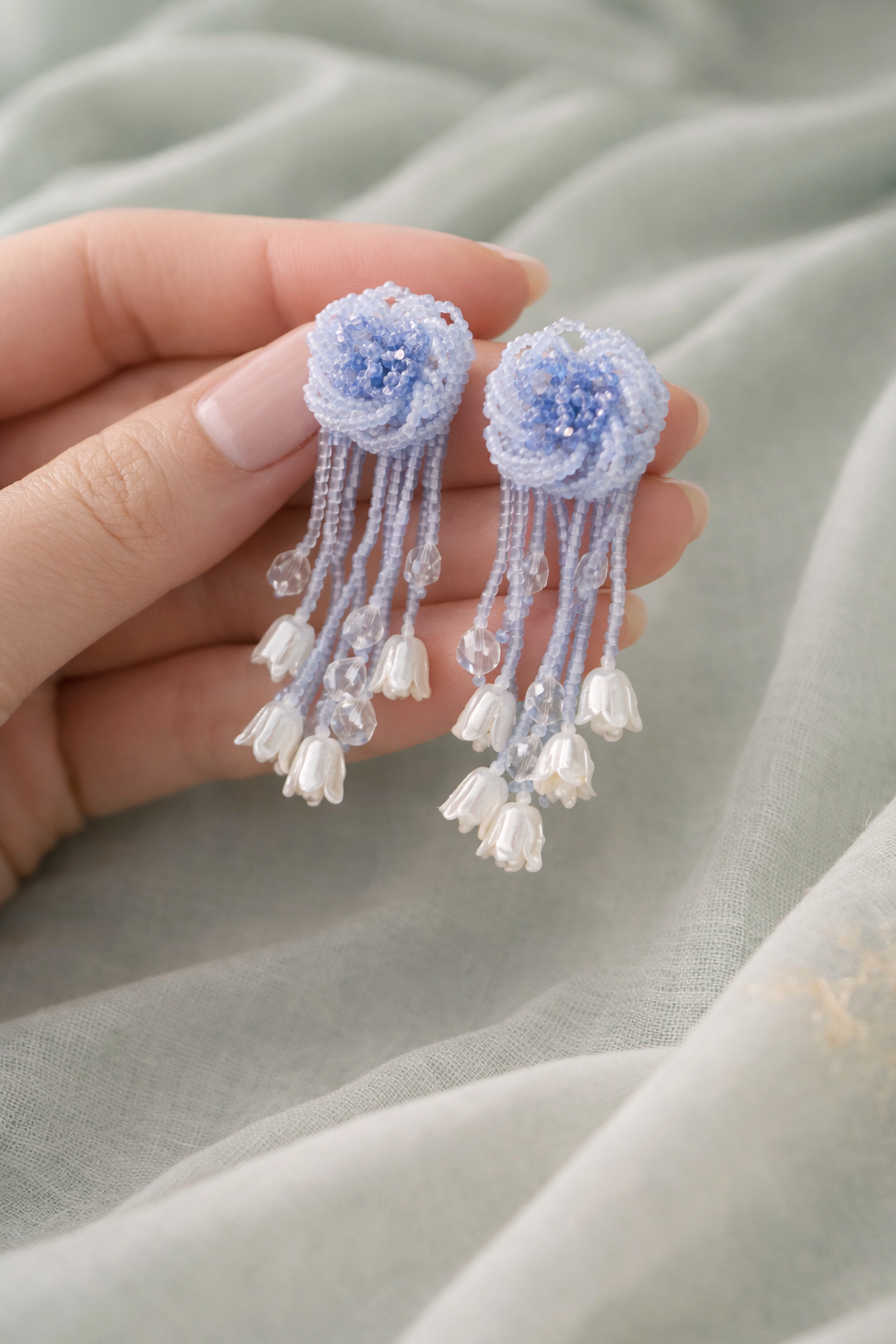 Anti-Tarnish, Ashaarya, Ashaarya Jewels, Ashaarya Jewellery, Hand holding a pair of delicate blue flower earrings with bead embellishments on a light fabric background.