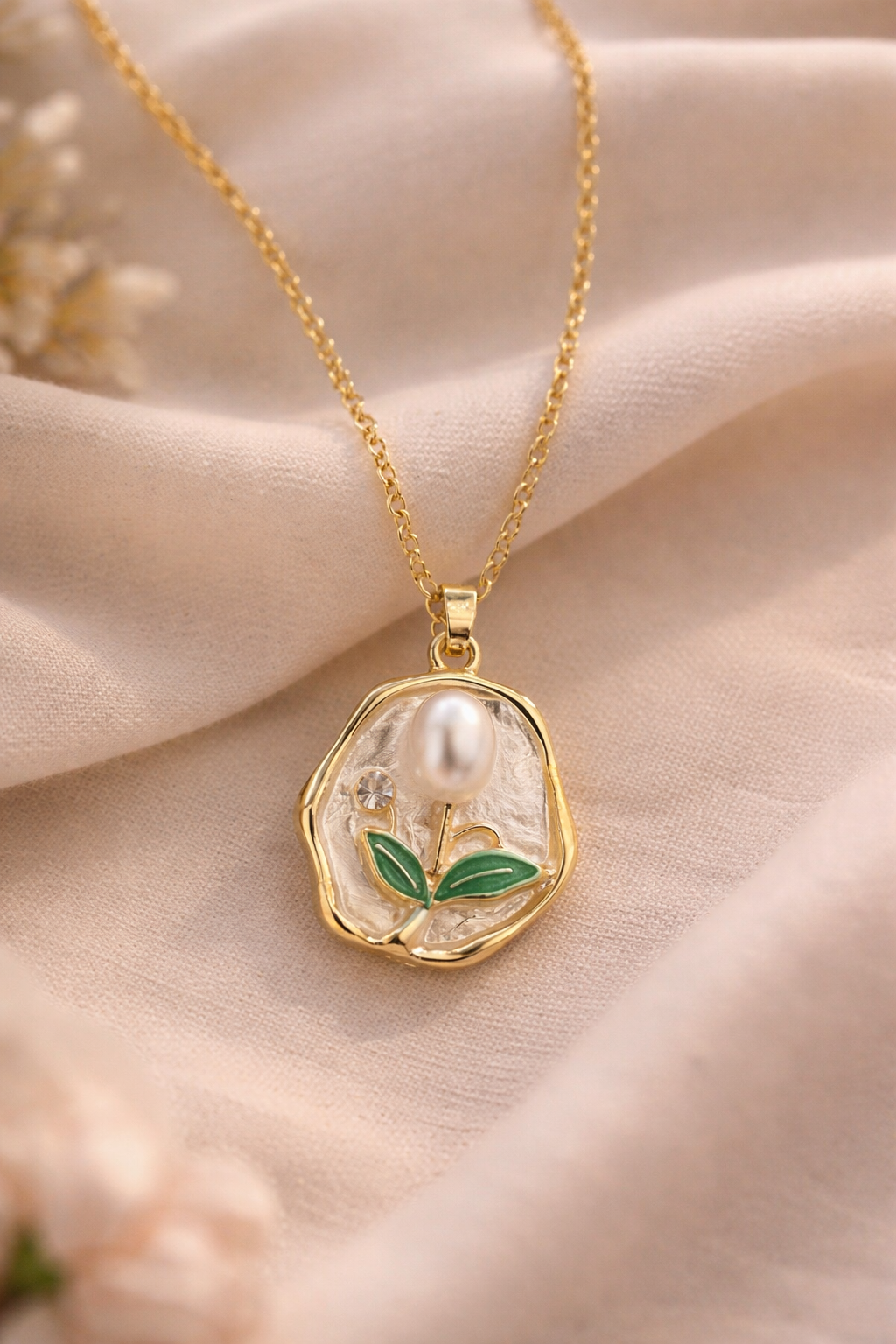 Anti-Tarnish, Ashaarya, Ashaarya Jewels, Ashaarya Jewellery, Gold necklace with a pendant featuring a pearl and green leaves on a soft beige fabric background.