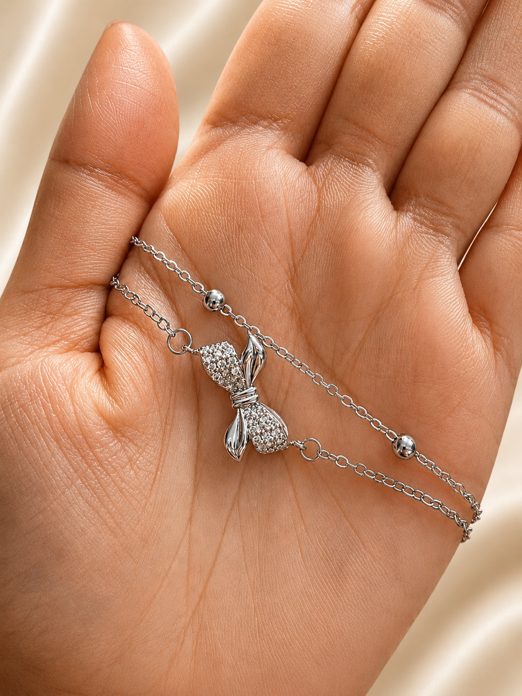 A silver-toned double-strand chain bracelet with a textured bow charm and small bead accents, displayed on an open hand. Elegant silver bow bracelet for women by Ashaarya made from premium 316L stainless steel with a skin-friendly finish.