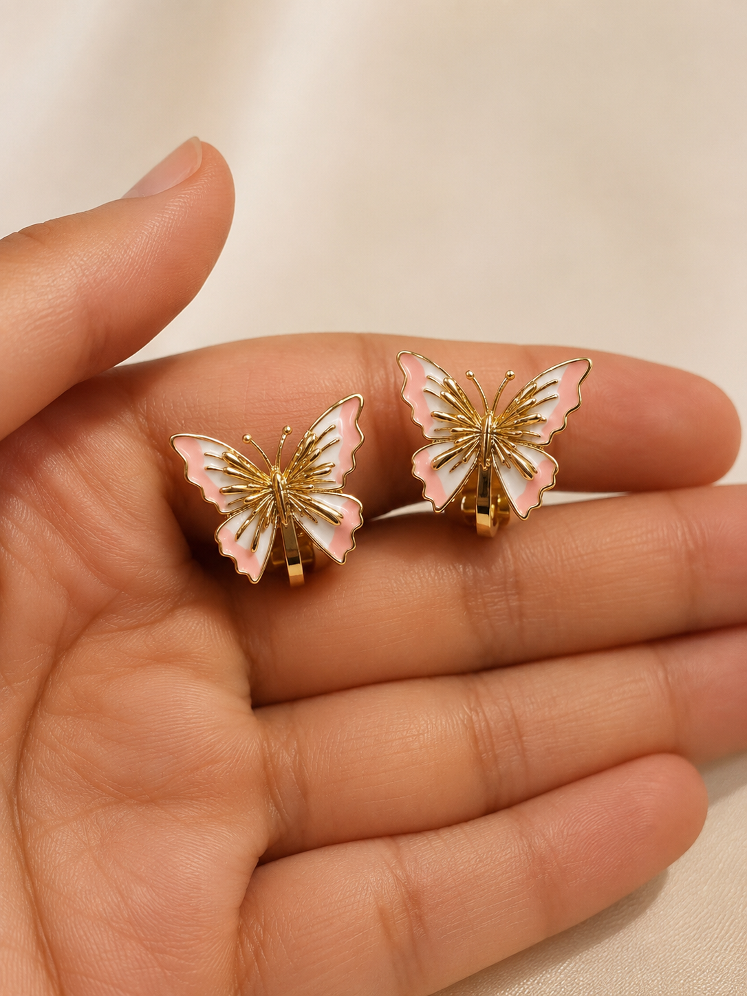 A pair of butterfly-shaped stud earrings with gold-tone metal and pale pink and white enamel, displayed on an open hand. Anti-tarnish gold butterfly earrings with pink enamel detailing