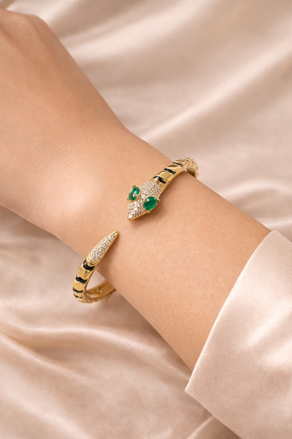 anti-tarnish comfort jewellery snake bracelet for women with American diamonds and emerald stones in 18kt gold tone luxury bracelet. Gold bracelet with green gemstones on a person's wrist against a satin background