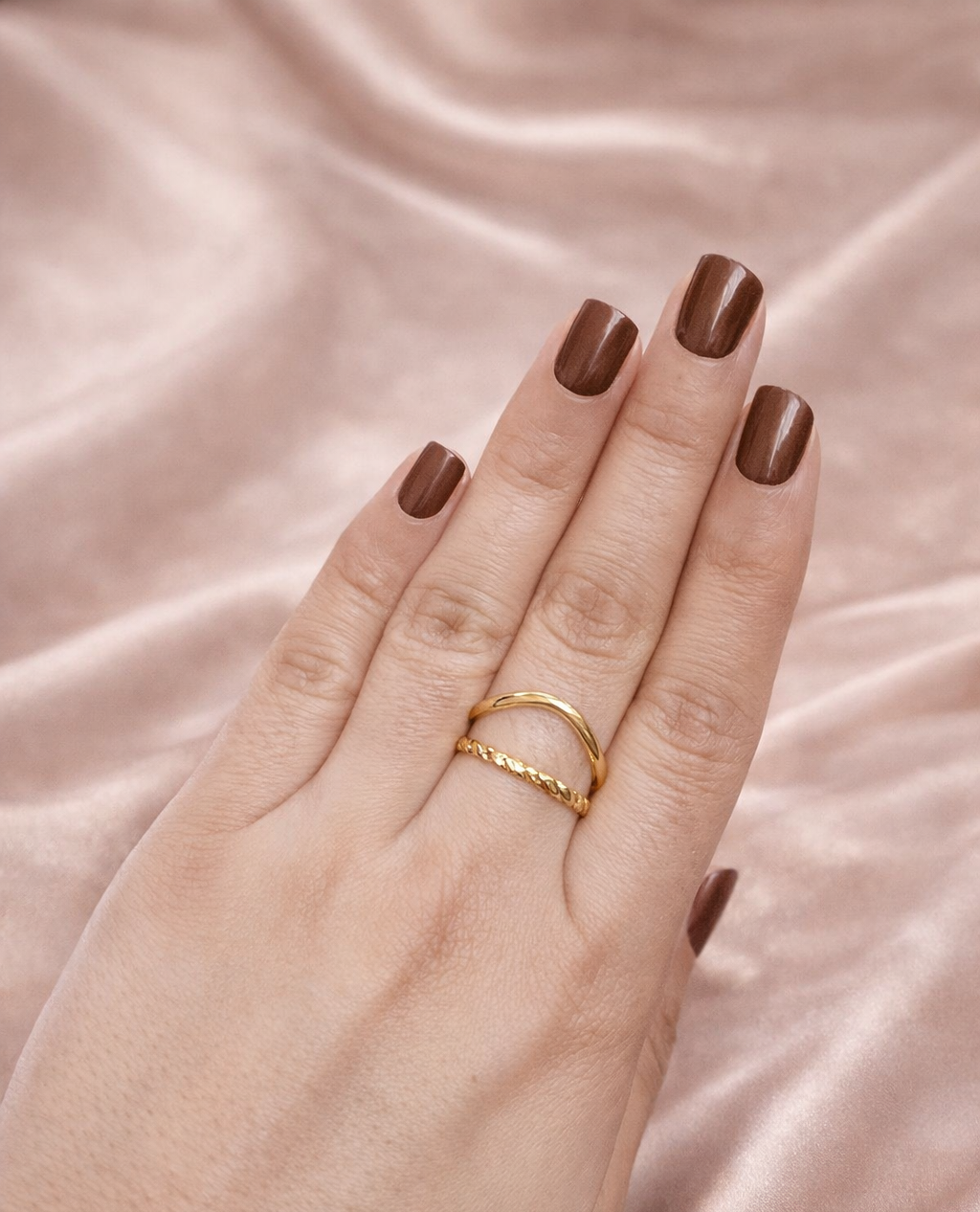 Anti-tarnish gold and silver duo ring stack worn on hand — adjustable comfort jewellery featuring twisted band and knot design, band rings for women, statement rings for women. Hand with gold ring on pink satin fabric