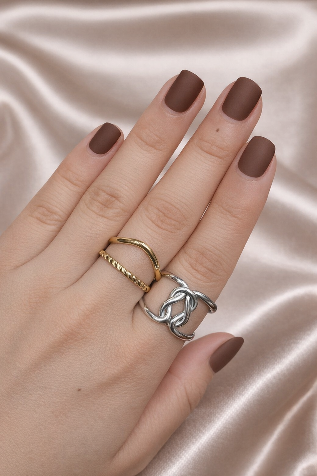 Anti-tarnish gold and silver duo ring stack worn on hand — adjustable comfort jewellery featuring twisted band and knot design, band rings for women, statement rings for women. Hand with brown nail polish wearing two rings on a satin background