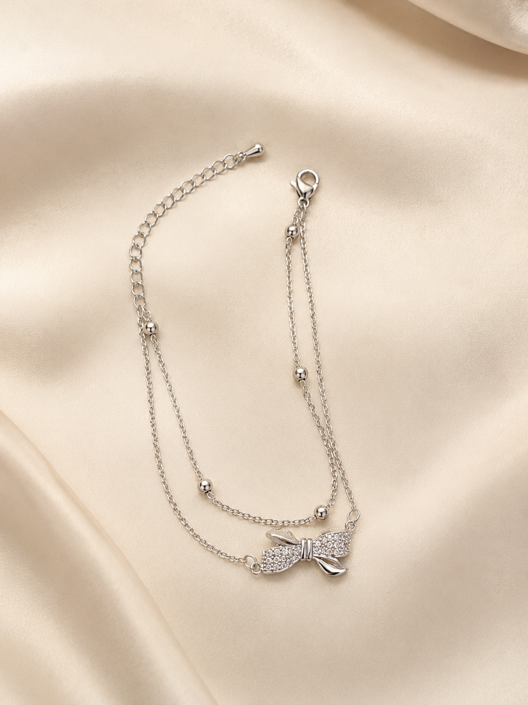 A delicate silver double-strand chain anklet featuring a small bow charm encrusted with clear stones, displayed on a draped cream satin fabric background. Bow of Grace Bracelet by Ashaarya – anti-tarnish silver bow bracelet with double chain and bead details Anti-tarnish Grace Bow Comfort Bracelet by Ashaarya crafted from 316L stainless steel with sparkling American Diamonds.