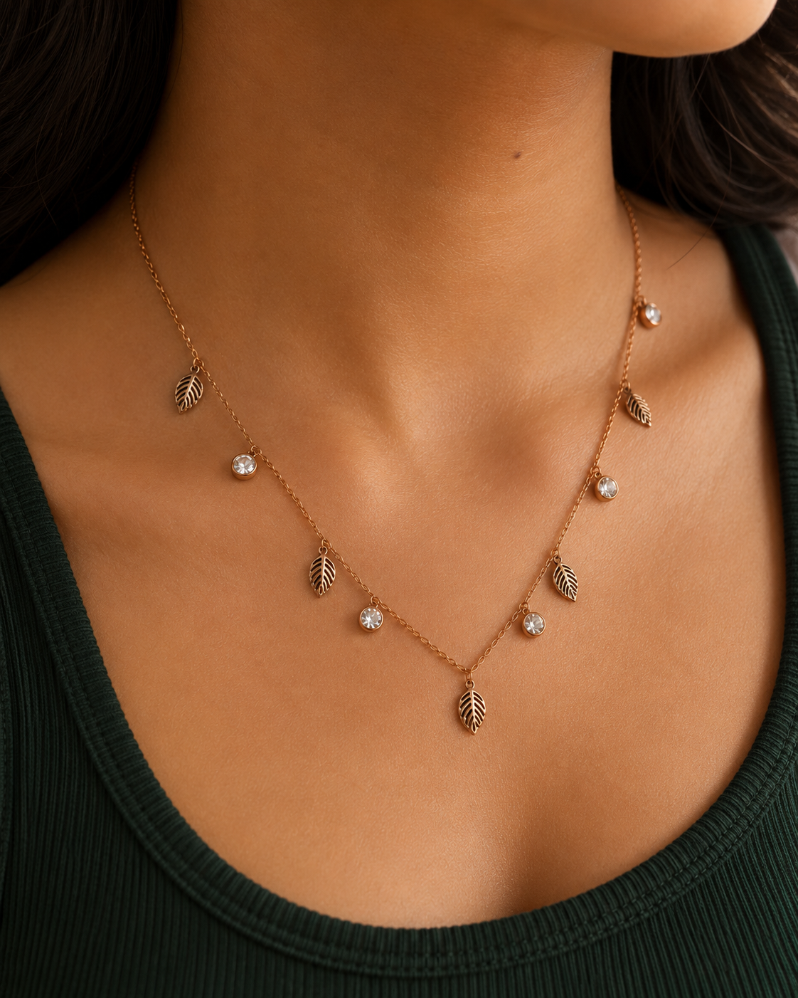 A close-up of a model's neck and chest wearing a delicate rose gold necklace with leaf and crystal charms over a dark green ribbed top. rose gold necklace for women, anti tarnish necklace, leaf pendant necklace, nature inspired necklace, lightweight necklace for women, stainless steel necklace, 18kt rose gold necklace, dainty necklace for women, everyday necklace, minimalist jewellery, comfort jewellery, waterproof necklace, skin friendly necklace, Ashaarya necklace