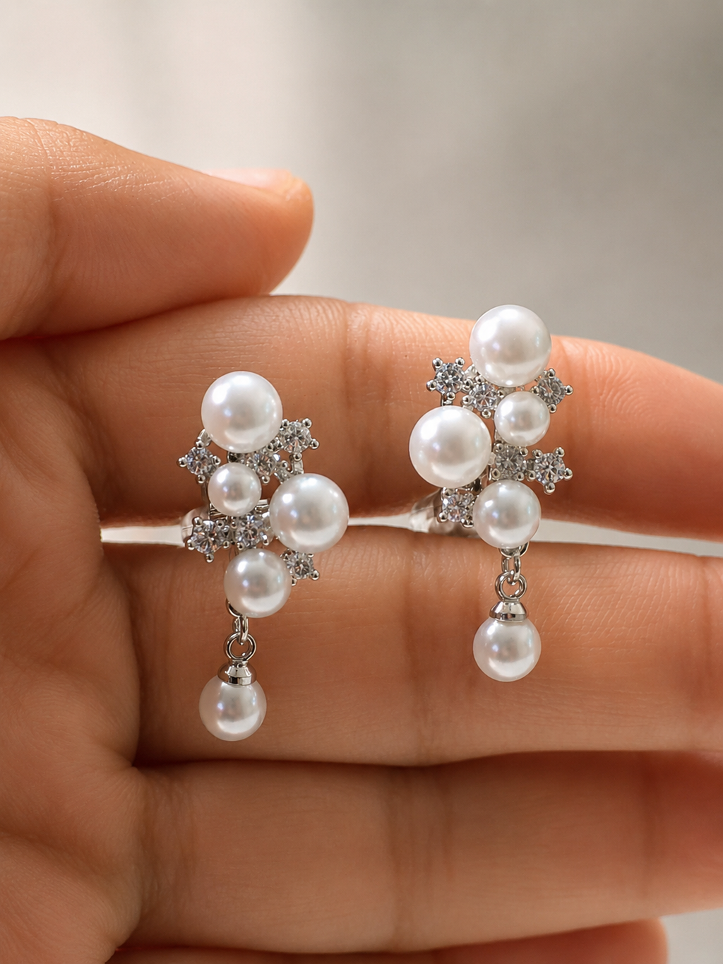 A pair of silver-toned drop earrings with white faux pearls and clear rhinestone accents, shown resting on a person's fingers.