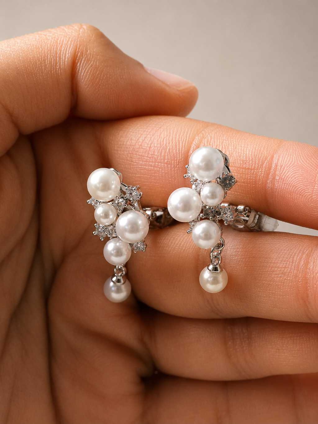 A pair of silver-toned drop earrings with clusters of white pearls and small clear stones, shown resting on a person's fingers against a soft beige background. Anti-tarnish pearl clip-on earrings for women with elegant crystal detailing