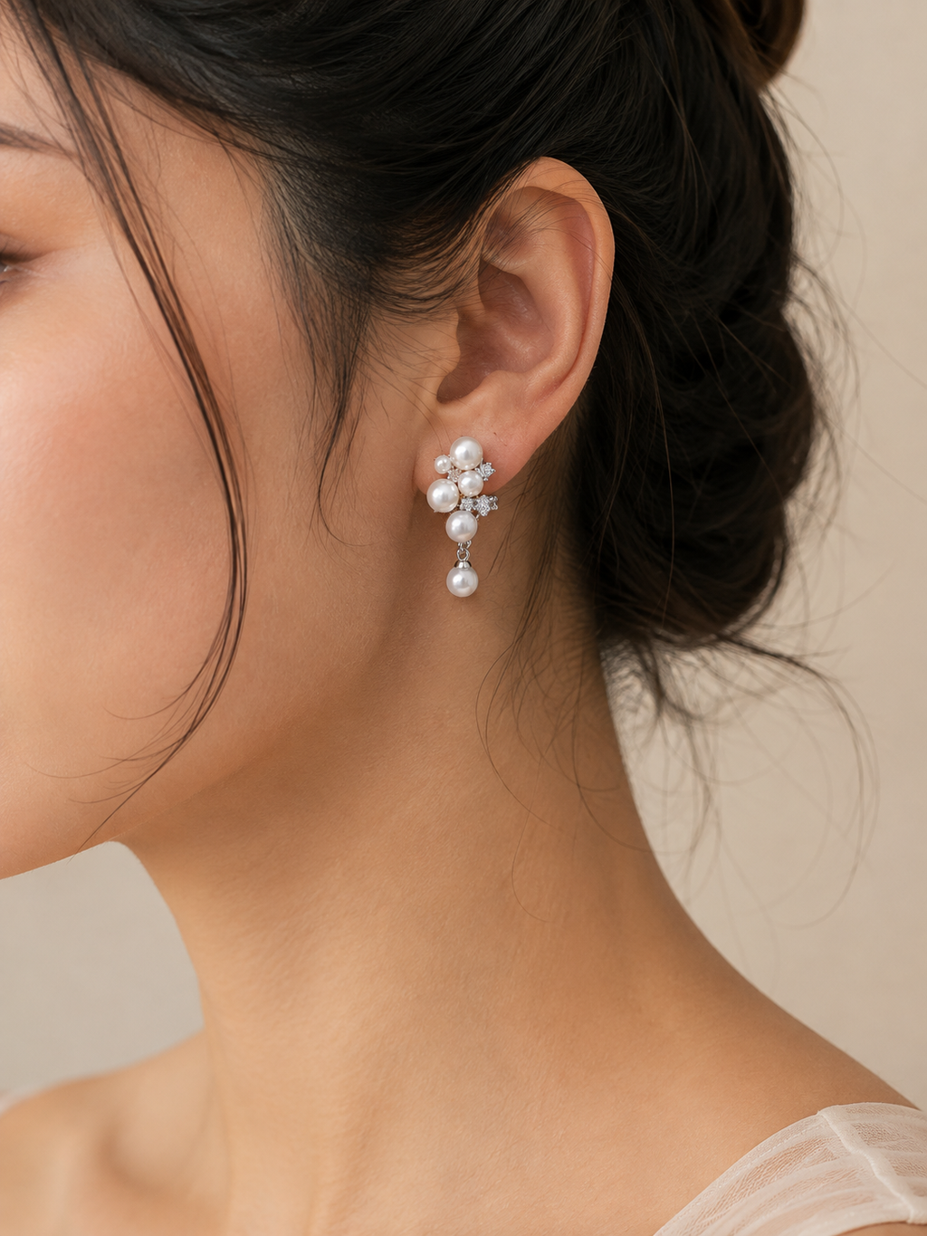 A close-up of a woman's ear wearing a delicate pearl and crystal dangle earring, with her dark hair pulled back and a soft neutral background.