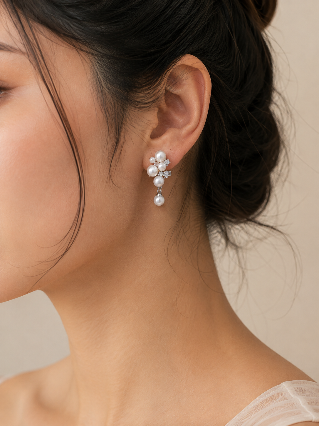 A close-up of a woman's ear wearing a delicate pearl and crystal dangle earring, with her dark hair pulled back and a soft neutral background.
