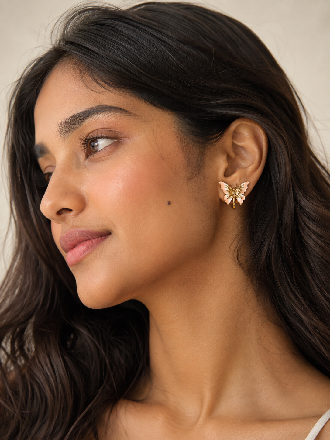 A close-up of a woman wearing a small gold butterfly-shaped stud earring, shown in profile against a soft beige background.