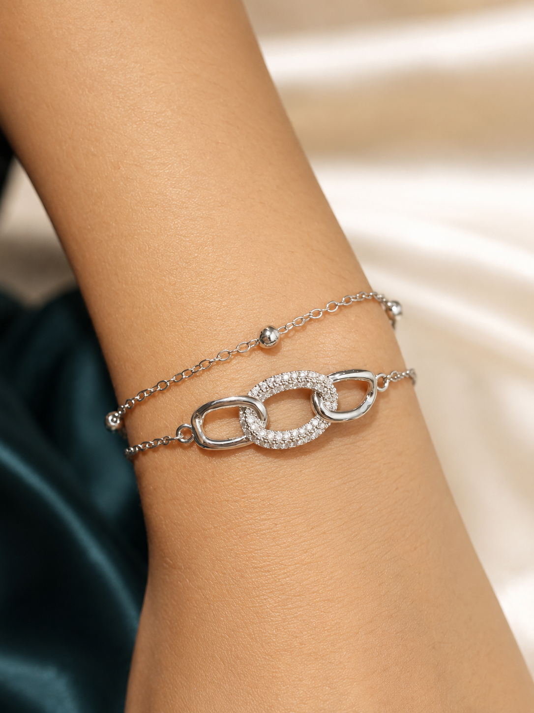 A close-up of a person's wrist wearing a delicate silver double-layer bracelet. The top layer features a fine chain with small silver beads, while the bottom layer has a larger chain with a central interlocking oval link paved with clear crystals. The Linked Grace Bracelet by Ashaarya, an anti-tarnish silver bracelet crafted in hypoallergenic 316L stainless steel with sparkling cubic zirconia links for lightweight everyday comfort.