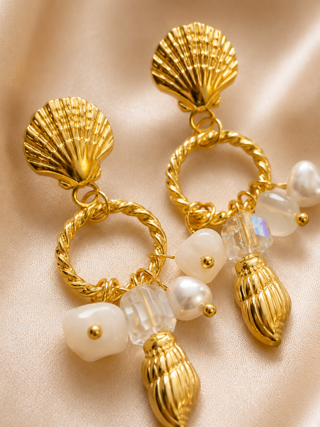 A pair of gold-toned statement earrings with scalloped shell-shaped studs, twisted rope rings, faux pearl beads, and small iridescent crystal drops, displayed on a soft beige satin fabric background. statement earrings for women, shell earrings, pearl drop earrings, beach inspired earrings, gold statement earrings, anti tarnish earrings, waterproof earrings, 18kt gold plated earrings, stainless steel earrings, coastal jewellery, lightweight earrings, comfort jewellery, vacation earrings, luxury earrings for