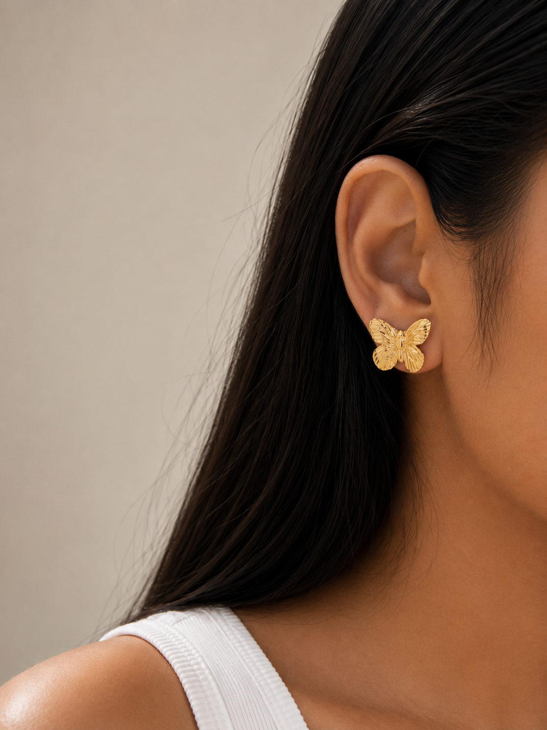 A close-up of a model's ear wearing a small textured gold butterfly stud earring, with her dark hair and white ribbed tank top visible against a plain beige background.Close-up of Golden Flutter Butterfly Stud Earrings by Ashaarya featuring intricate butterfly wing detailing.