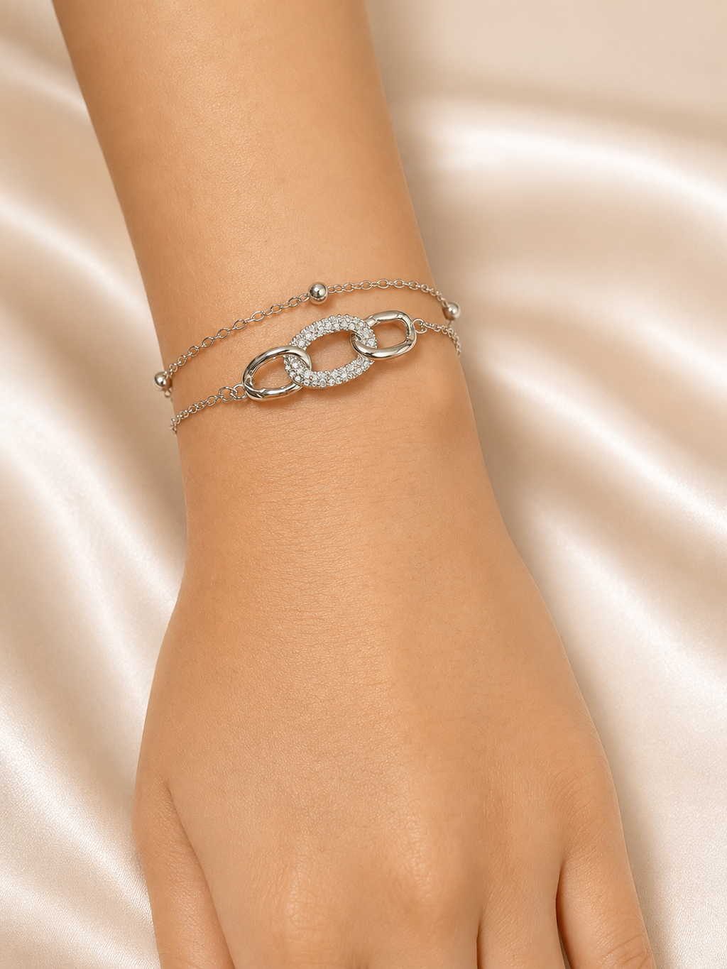 A close-up of a person's wrist wearing two delicate silver-tone chain bracelets, featuring a central interlocking oval link design paved with small clear stones, set against a soft beige satin fabric background.