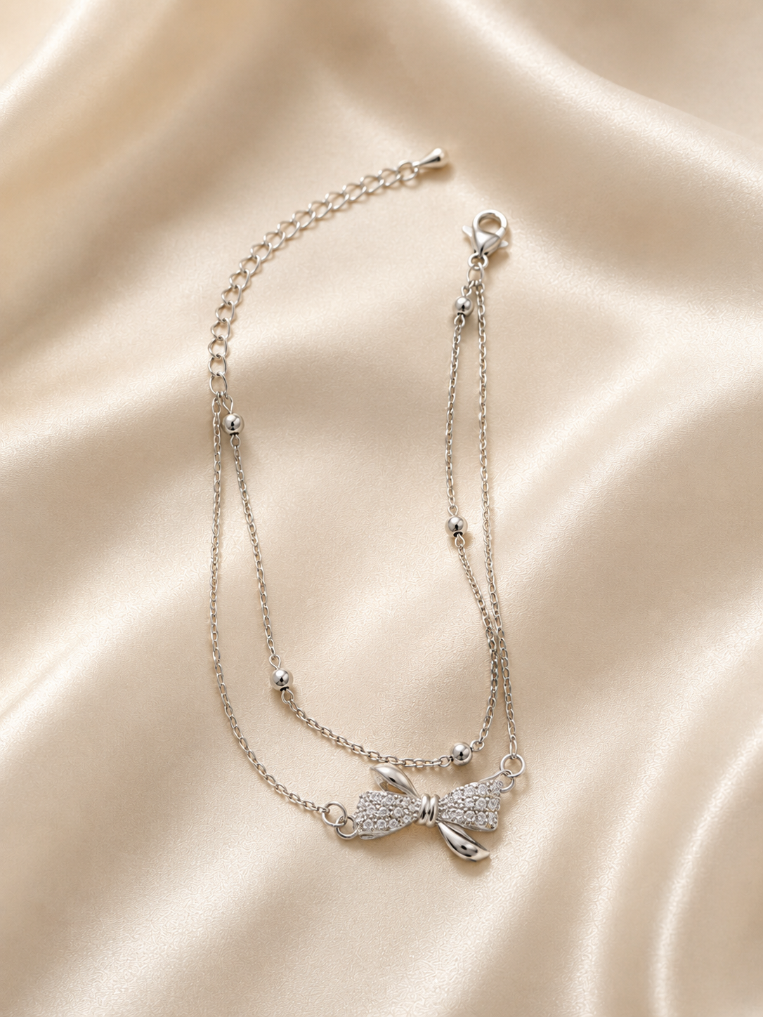 A silver double-strand necklace with a small bow charm covered in clear rhinestones, displayed on a draped beige satin fabric background. Grace Bow Comfort Bracelet by Ashaarya showcasing a delicate bow motif with polished bead accents and an adjustable chain.