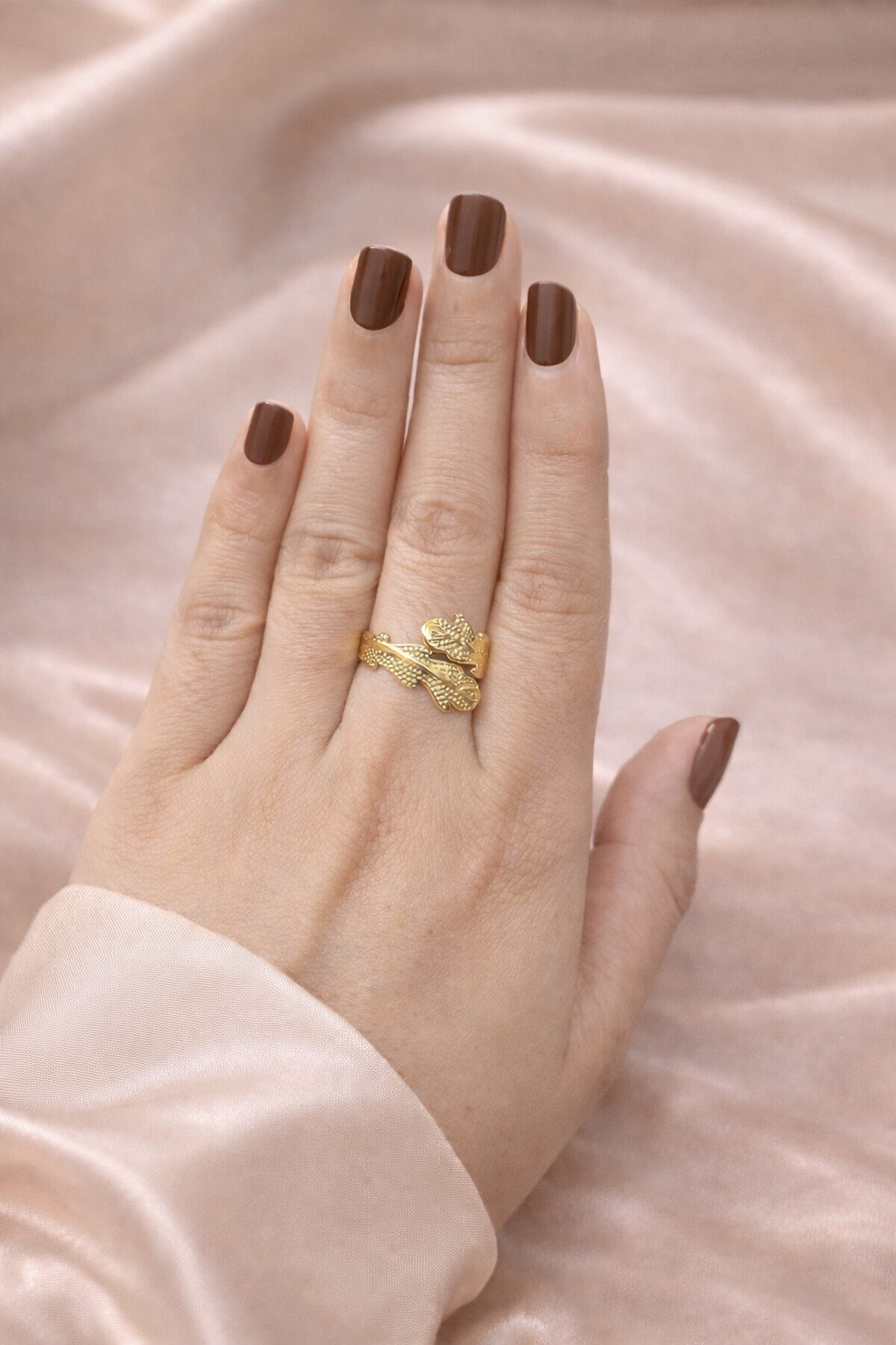Anti-tarnish gold leaf design ring worn on hand, featuring textured wrap detailing, photographed against champagne satin background — comfort jewellery by Ashaarya. Hand wearing a gold ring with brown nail polish on a beige fabric background