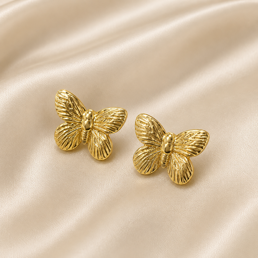 A pair of gold butterfly-shaped stud earrings with a textured, ribbed wing pattern, displayed on a draped beige satin fabric background.