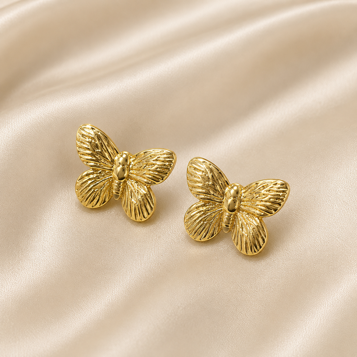 A pair of gold butterfly-shaped stud earrings with a textured, ribbed wing pattern, displayed on a draped beige satin fabric background.