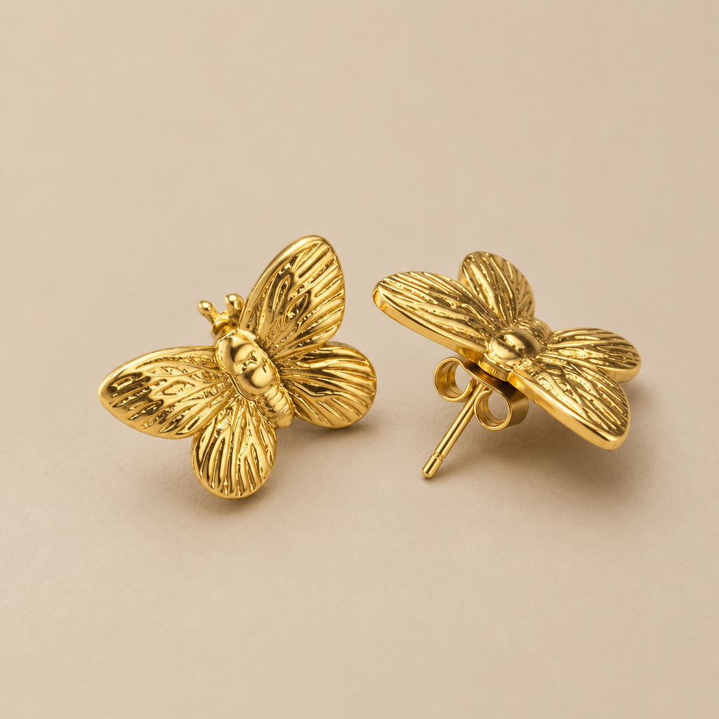 A pair of gold butterfly stud earrings with textured wings, shown on a plain beige background. One earring is facing forward to show the butterfly detail, and the other is turned to show the post and backing. Golden Flutter Butterfly Stud Earrings by Ashaarya in premium 18KT gold tone with textured butterfly wings.