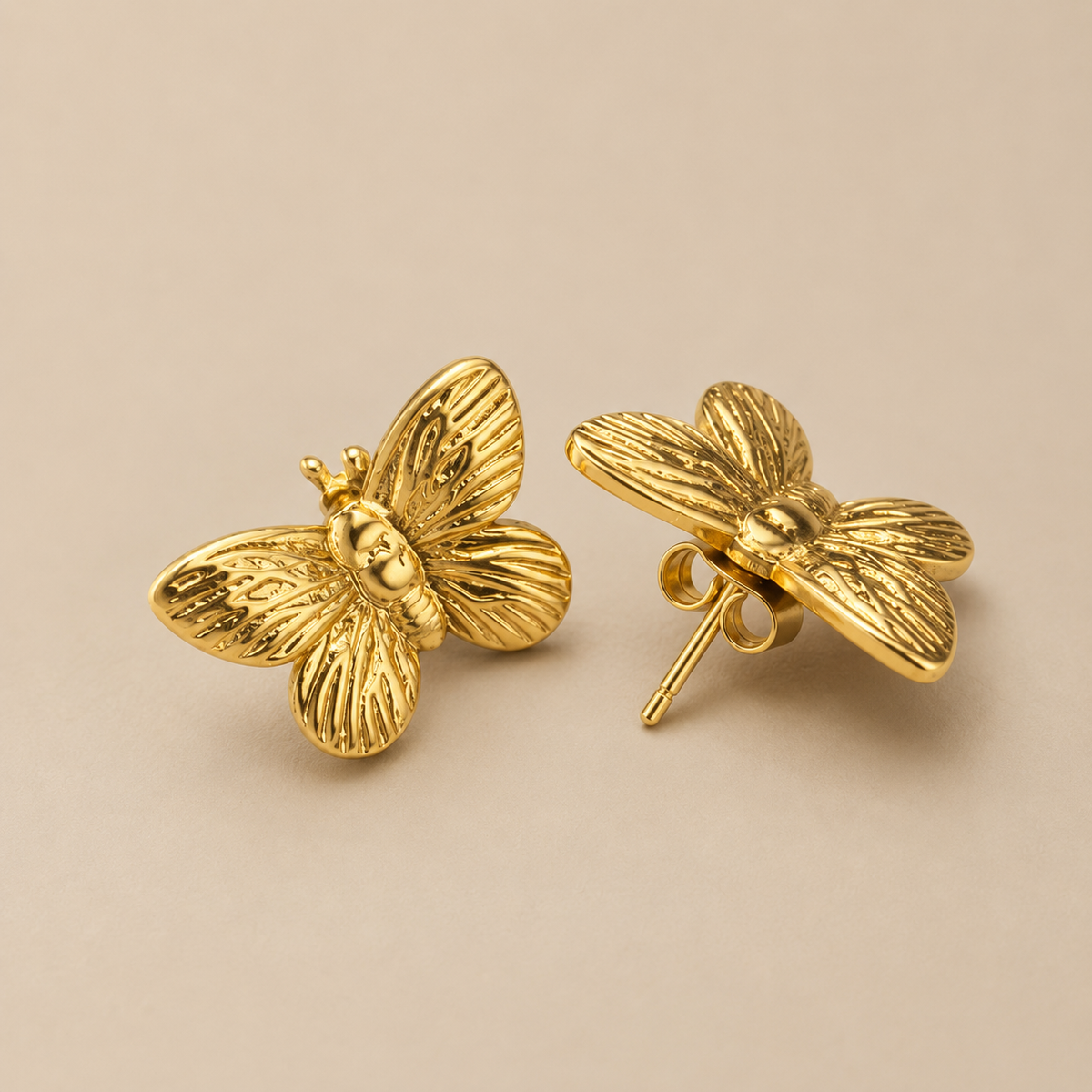 A pair of gold butterfly stud earrings with textured wings, shown on a plain beige background. One earring is facing forward to show the butterfly detail, and the other is turned to show the post and backing. Golden Flutter Butterfly Stud Earrings by Ashaarya in premium 18KT gold tone with textured butterfly wings.