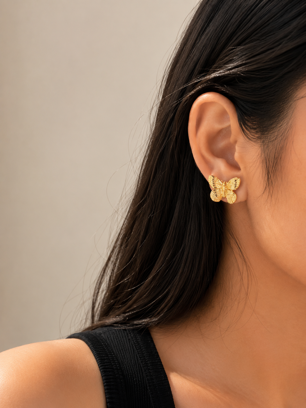 A close-up of a woman's ear wearing a small gold butterfly stud earring, with her dark hair and black top visible against a soft neutral background.  Model wearing Golden Flutter Butterfly Stud Earrings by Ashaarya with textured gold butterfly design.