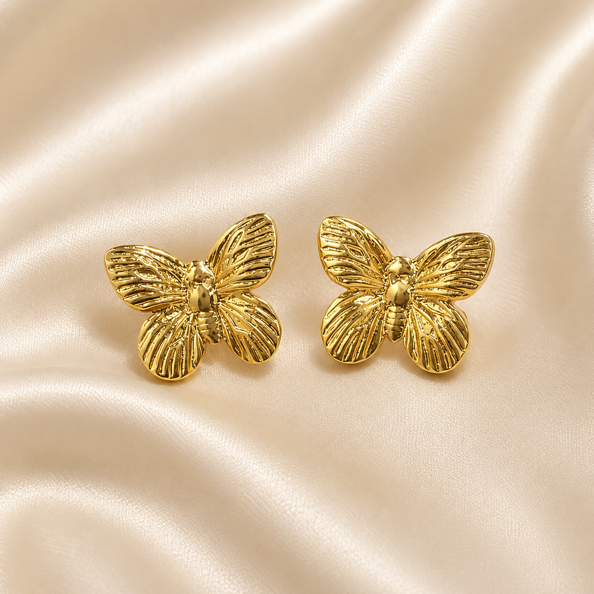 A pair of gold butterfly-shaped stud earrings displayed on a draped cream satin fabric background. Golden Flutter Butterfly Stud Earrings by Ashaarya in premium 18KT gold tone with textured butterfly wings.
