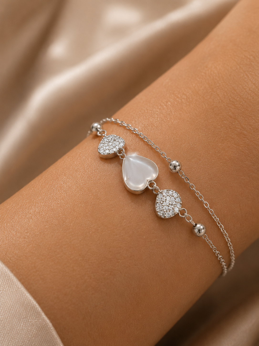 A delicate silver chain bracelet with three heart charms, including a central milky white heart and two pavé-set heart charms, shown worn on a wrist.