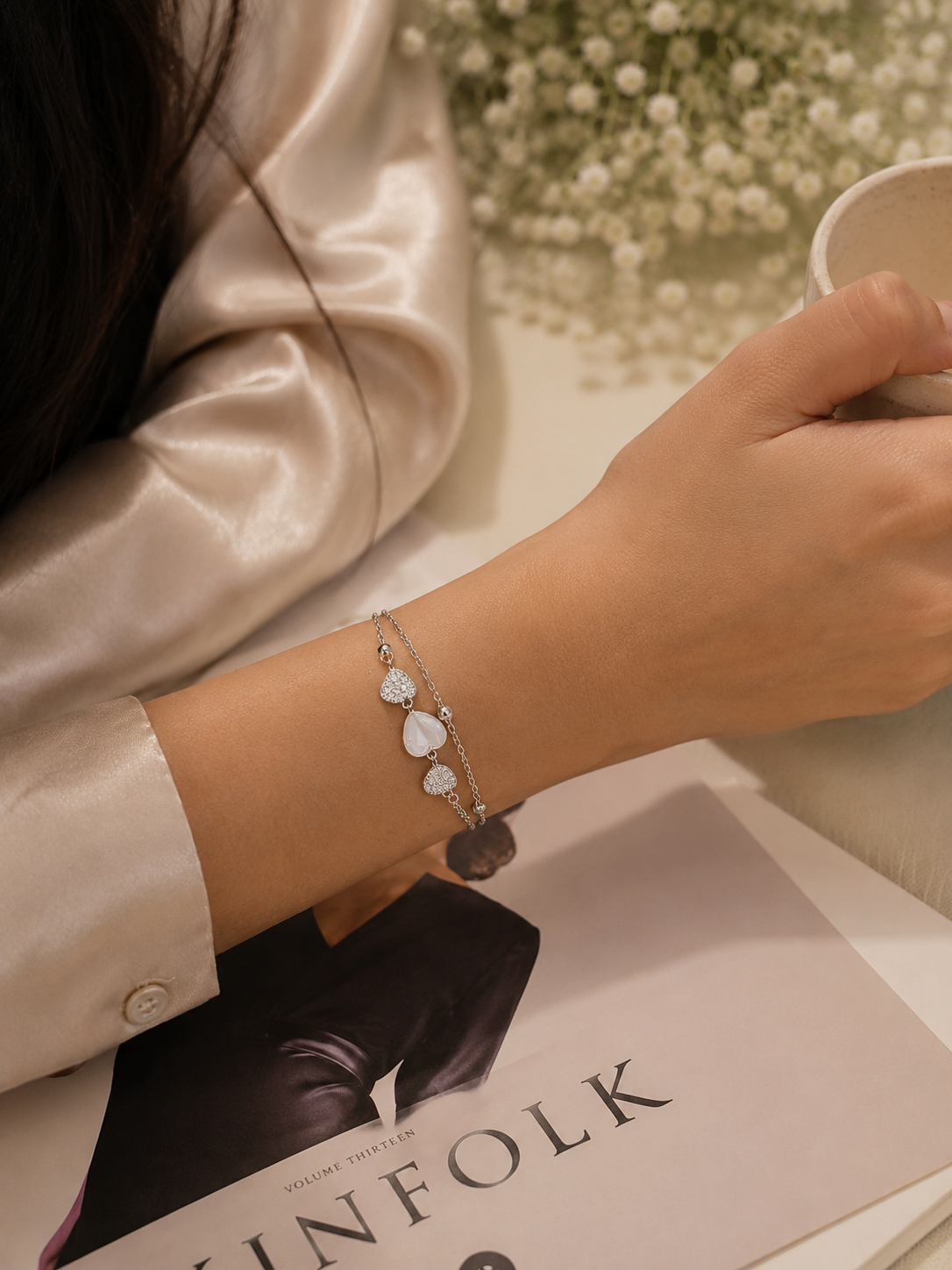 A delicate silver-toned chain bracelet with three white heart-shaped charms and a dark gray stone pendant, worn on a person's wrist. The person is wearing a champagne-colored satin shirt and holding a white ceramic cup, with a magazine and baby's breath flowers in the background.