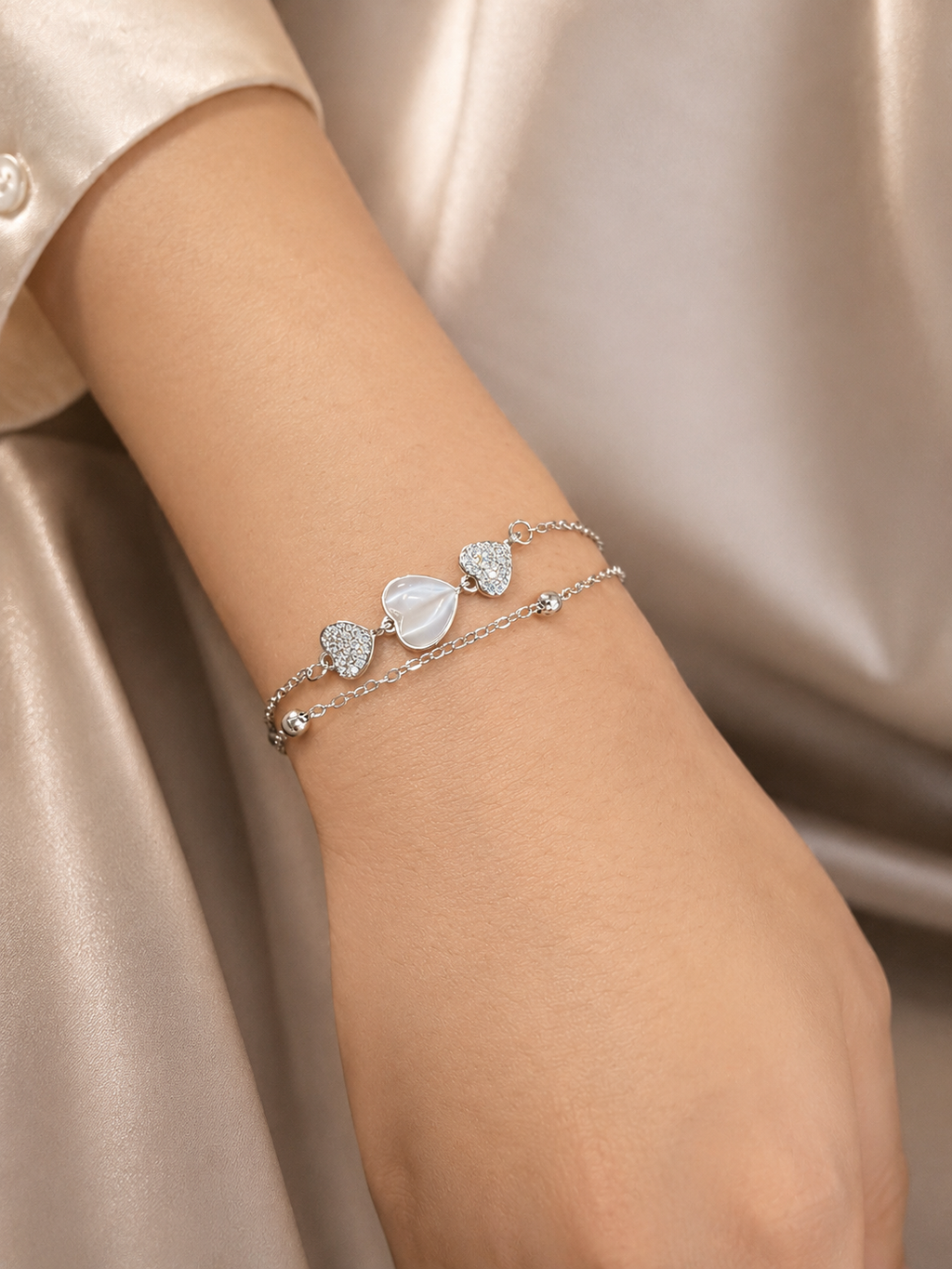 A close-up of a person's wrist wearing a delicate silver double-strand bracelet with three heart-shaped charms, including one mother-of-pearl heart and two pavé-set heart charms, against a champagne satin background. Model wearing the Heartstrings Bracelet by Ashaarya, a lightweight anti-tarnish silver bracelet with a mother-of-pearl heart and American Diamonds.