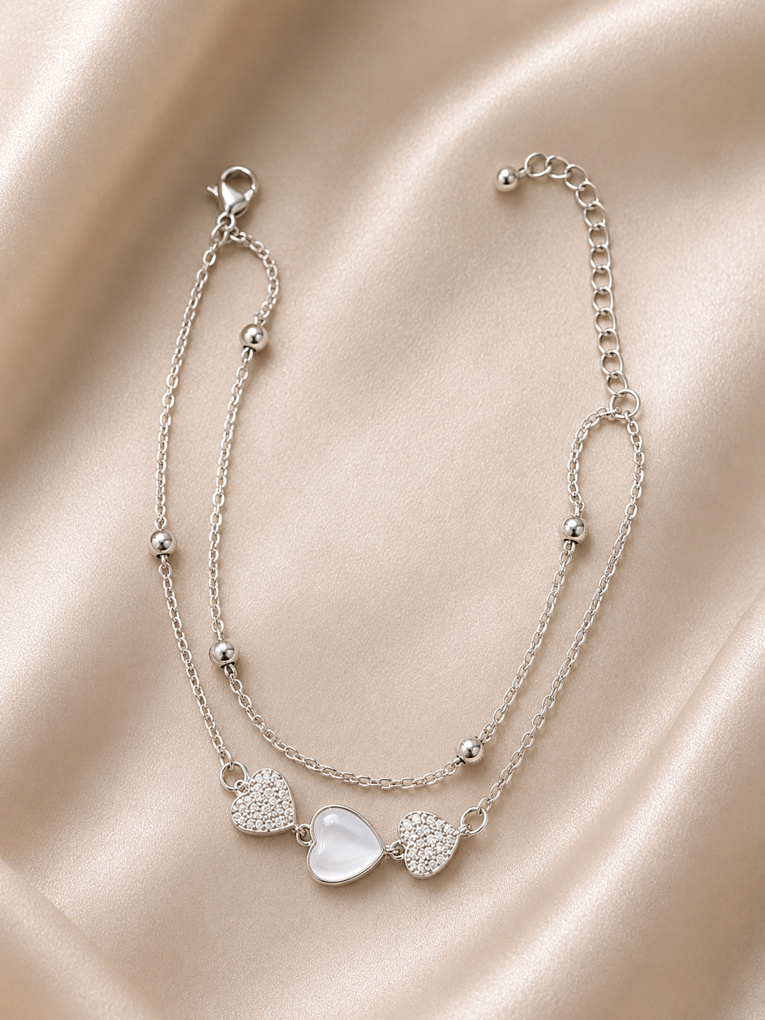 A delicate silver double-layered anklet featuring three heart charms, one of which is a smooth white oval stone, displayed on a draped champagne satin fabric.