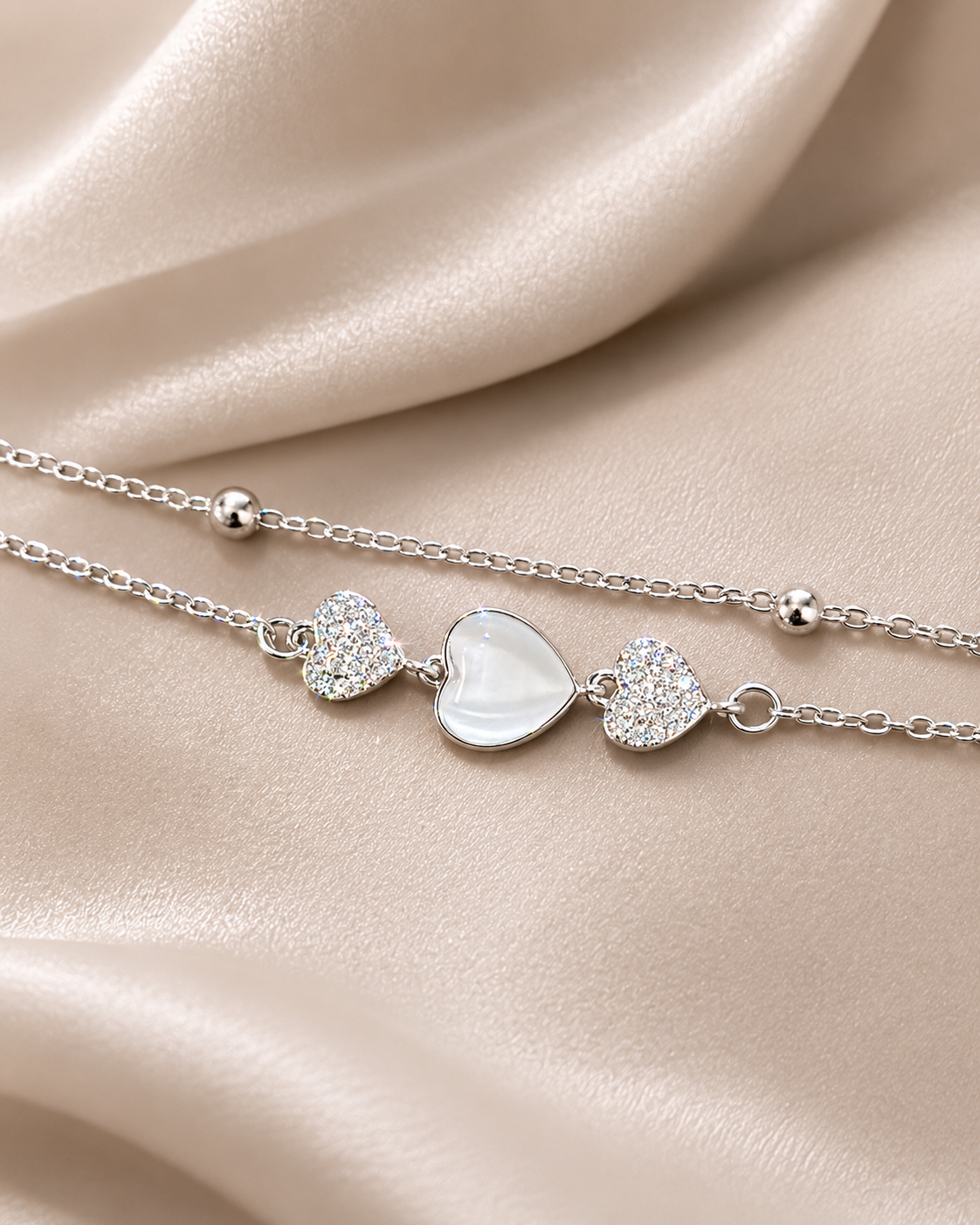 A delicate silver chain necklace with three heart charms, featuring a central white mother-of-pearl heart flanked by two clear crystal-encrusted hearts, displayed on a draped beige satin fabric. Heartstrings Bracelet by Ashaarya featuring a mother-of-pearl heart, American Diamond accents and a lightweight anti-tarnish silver bracelet for women.