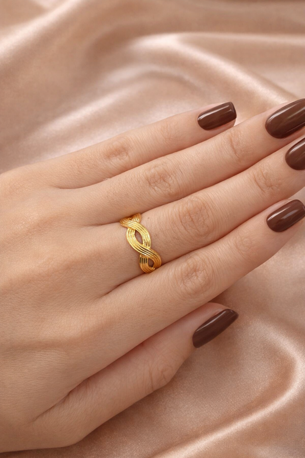 Anti-tarnish gold adjustable twist ring with infinity rope design, open back for flexible sizing, displayed on hand against satin background — lightweight comfort jewellery by Ashaarya, Gold ring on a hand with brown nail polish against a beige fabric background