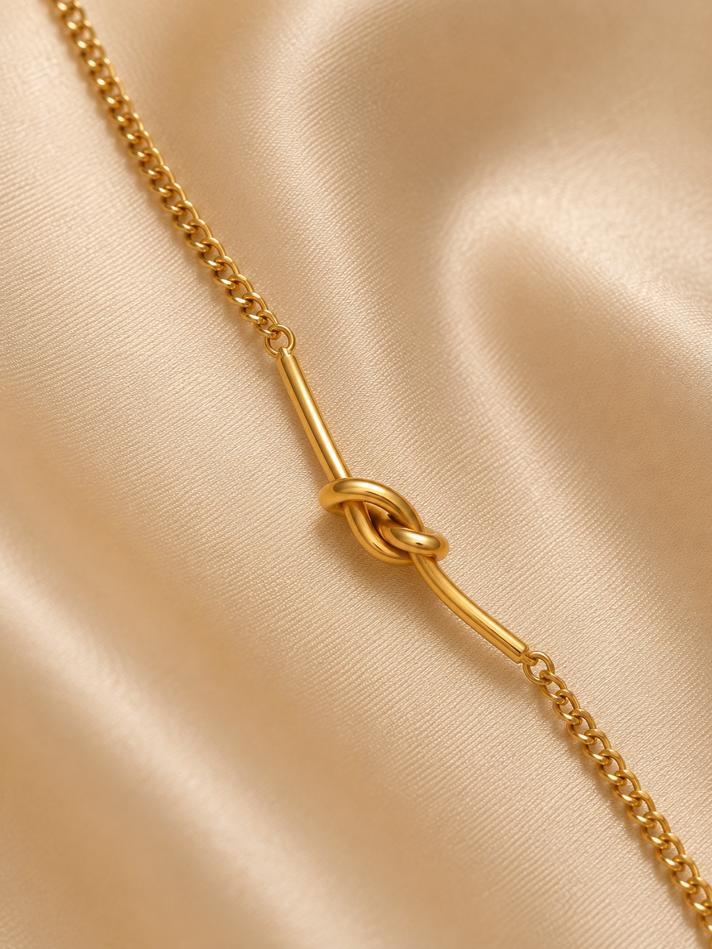 A gold chain necklace featuring a central knotted bar detail, displayed on a draped beige satin fabric background.