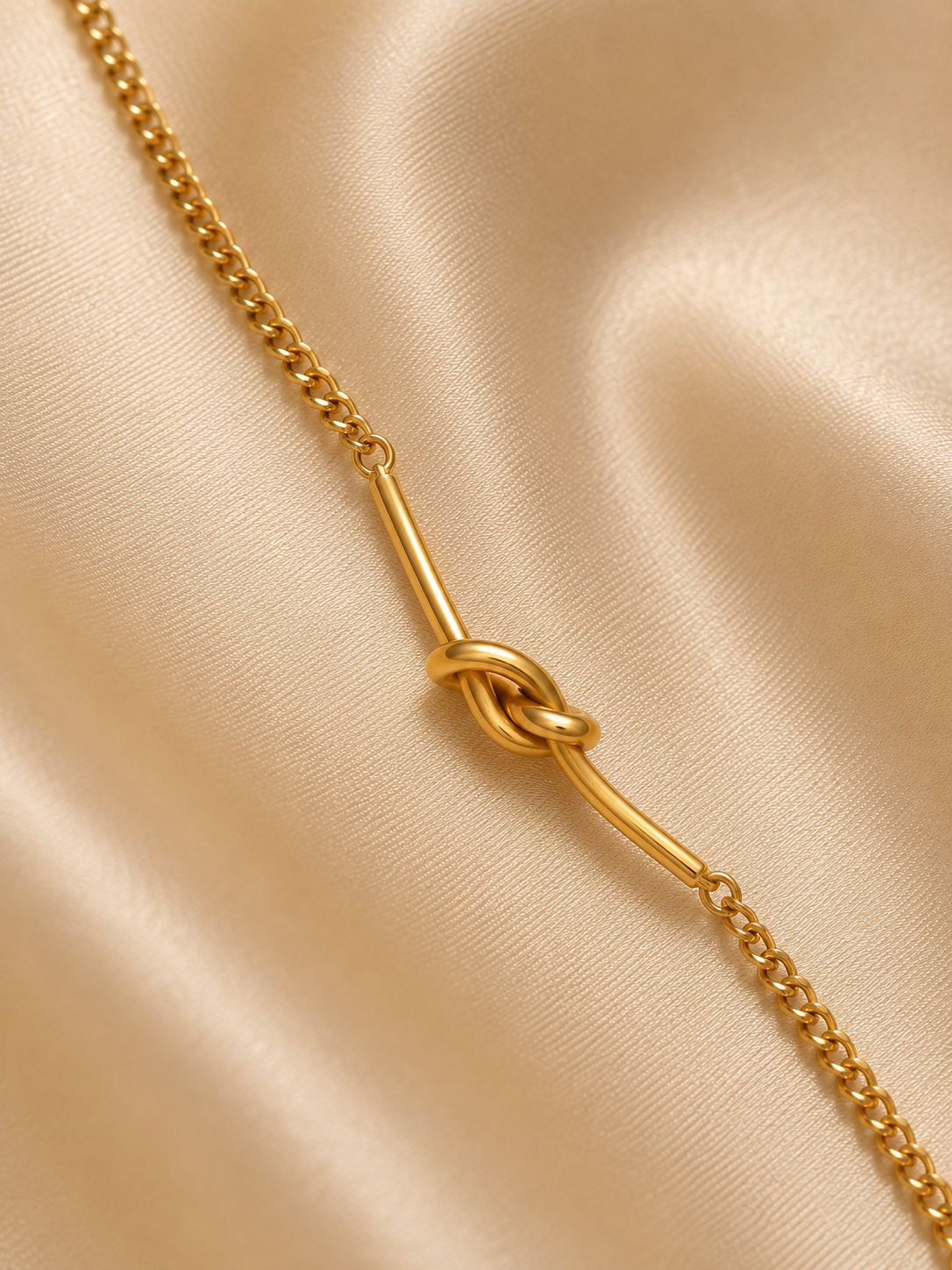A gold chain necklace featuring a central knotted bar detail, displayed on a draped beige satin fabric background.