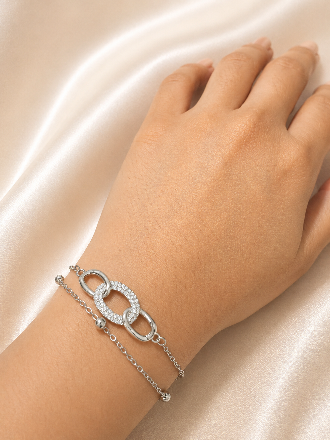 A silver-toned double-strand bracelet with a pavé-set oval link and a smaller chain link, shown worn on a hand against a draped champagne satin background. The Linked Grace Bracelet by Ashaarya, an anti-tarnish silver bracelet crafted in hypoallergenic 316L stainless steel with sparkling cubic zirconia links for lightweight everyday comfort.