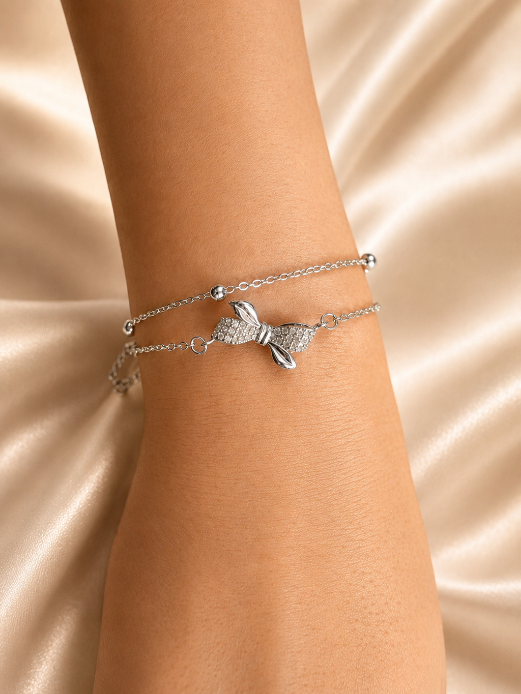 A close-up of a person's wrist wearing two delicate silver bracelets, including a bow-shaped charm bracelet and a thin chain bracelet, set against a draped champagne satin background. Lightweight silver bracelet with American Diamonds by Ashaarya designed for comfortable all-day wear.