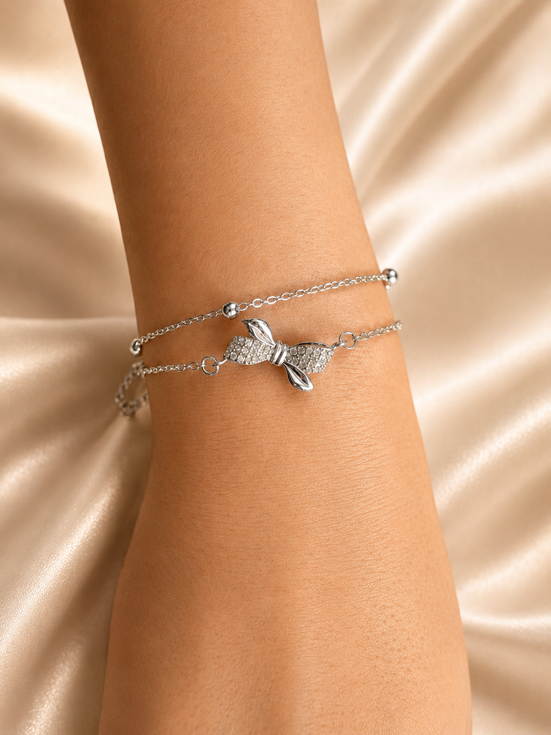 A close-up of a person's wrist wearing two delicate silver bracelets, including a bow-shaped charm bracelet and a thin chain bracelet, set against a draped champagne satin background. Lightweight silver bracelet with American Diamonds by Ashaarya designed for comfortable all-day wear.