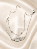 A silver double-layered chain bracelet with a central interlocking oval link design paved with small clear stones, displayed on a draped cream satin fabric background. The Linked Grace Bracelet by Ashaarya, an anti-tarnish silver bracelet crafted in hypoallergenic 316L stainless steel with sparkling cubic zirconia links for lightweight everyday comfort.