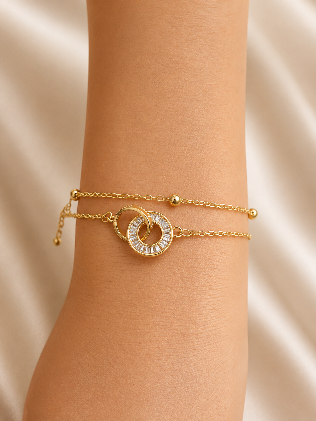 A close-up of a gold double-strand bracelet worn on a wrist, featuring a central interlocking circular design with a clear stone halo, set against a soft beige satin background. Linked in Light anti-tarnish gold bracelet for women with interlocking circular motif and baguette-cut stones by Ashaarya