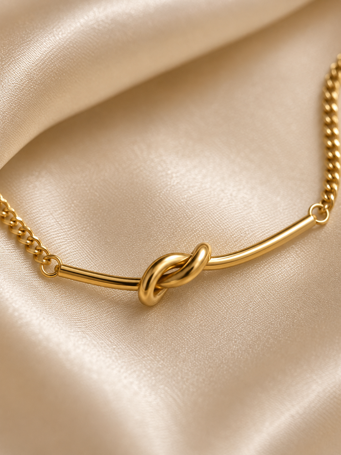 Minimal anti-tarnish gold knot bracelet displayed on a neutral satin background for a luxury jewellery aesthetic.. A gold-toned necklace with a central knotted bar detail and a chain, displayed on draped champagne satin fabric.
