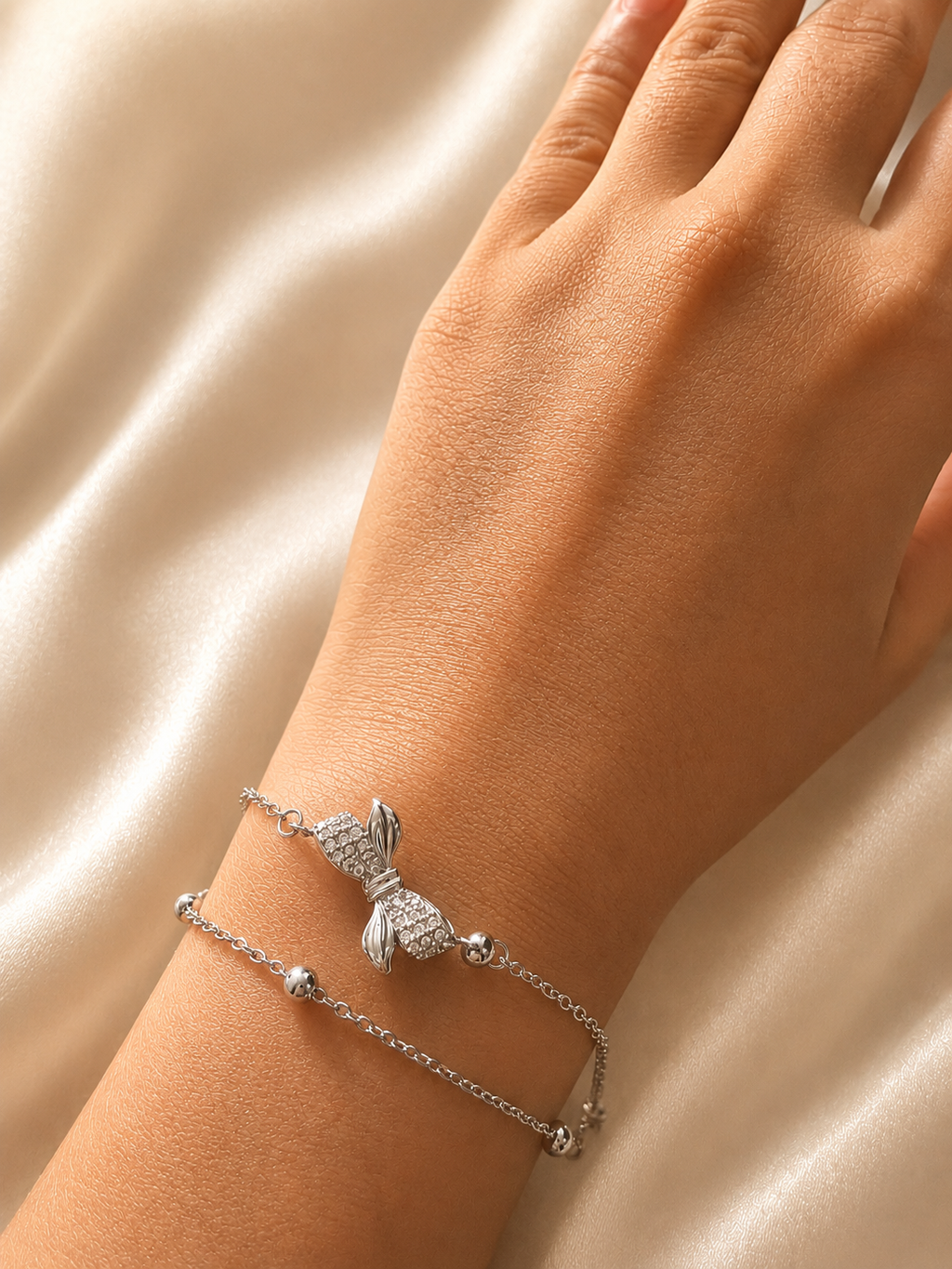 A close-up of a person's wrist wearing two delicate silver bracelets, including a bunny charm bracelet and a thin beaded chain bracelet, set against a soft beige satin background. Dainty everyday silver bracelet by Ashaarya with a crystal-studded bow centerpiece, anti-tarnish finish, and comfortable fit.