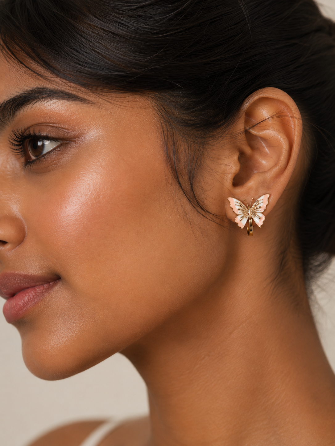 A close-up side profile of a woman wearing a small butterfly-shaped stud earring with white and pink enamel and gold-tone metal. Pink butterfly earrings for women with delicate gold detailing
