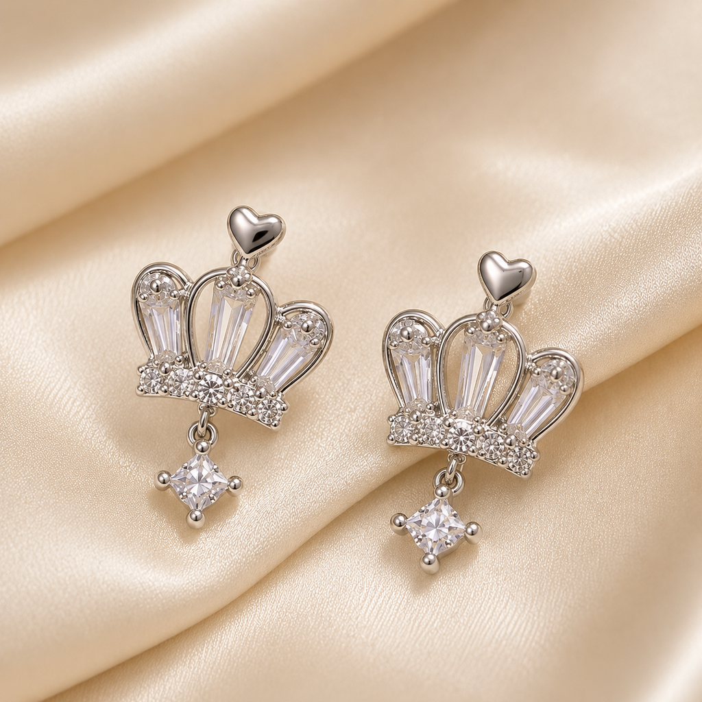 Crown-shaped earrings with heart and diamond details on a beige fabric background. royal trio earrings set, crown earrings for women, silver crown earrings, crown stud earrings, crown drop earrings, stud earrings for women, anti tarnish earrings, stainless steel earrings, lightweight earrings, skin friendly earrings, hypoallergenic earrings, cubic zirconia earrings, silver earrings for girls, everyday earrings, party wear earrings, elegant stud earrings, dainty earrings, premium earrings for women, earrings