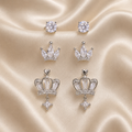 Set of silver crown-shaped earrings with clear gemstones on a beige satin background. royal trio earrings set, crown earrings for women, silver crown earrings, crown stud earrings, crown drop earrings, stud earrings for women, anti tarnish earrings, stainless steel earrings, lightweight earrings, skin friendly earrings, hypoallergenic earrings, cubic zirconia earrings, silver earrings for girls, everyday earrings, party wear earrings, elegant stud earrings, dainty earrings, premium earrings for women, earri