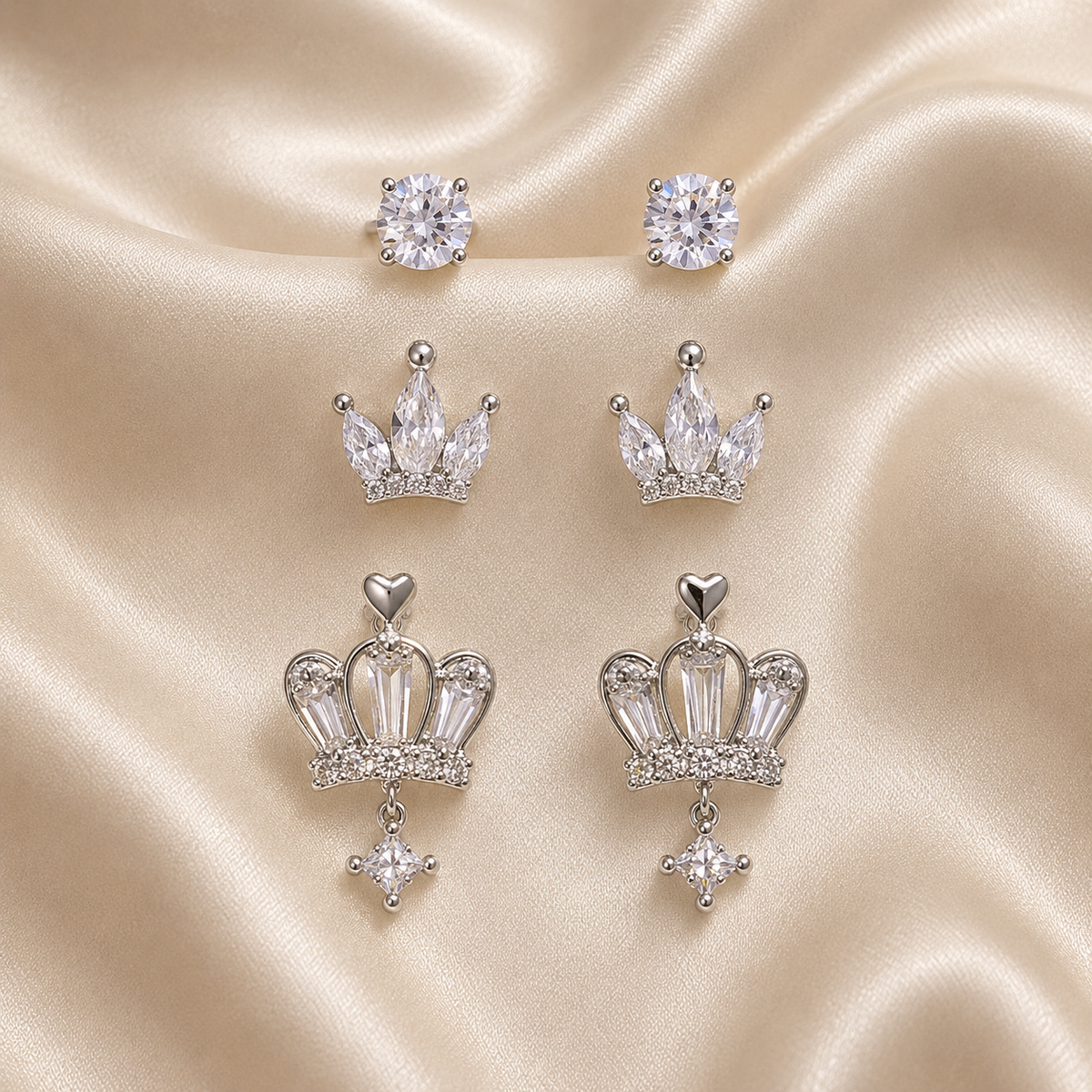 Set of silver crown-shaped earrings with clear gemstones on a beige satin background. royal trio earrings set, crown earrings for women, silver crown earrings, crown stud earrings, crown drop earrings, stud earrings for women, anti tarnish earrings, stainless steel earrings, lightweight earrings, skin friendly earrings, hypoallergenic earrings, cubic zirconia earrings, silver earrings for girls, everyday earrings, party wear earrings, elegant stud earrings, dainty earrings, premium earrings for women, earri