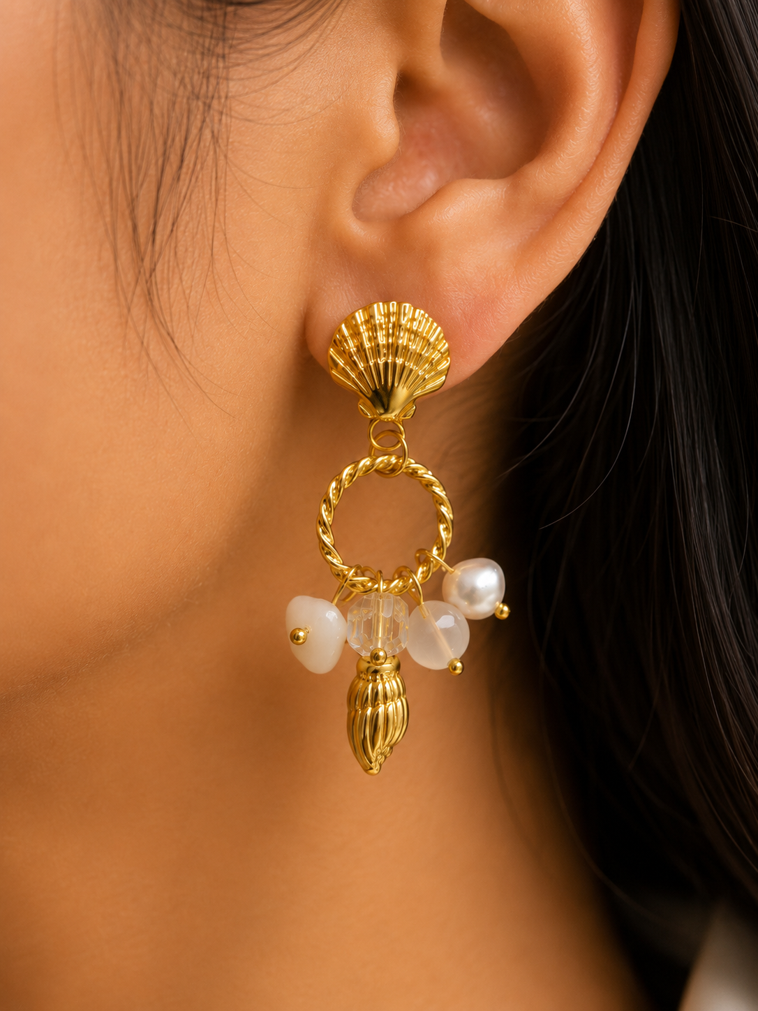 A close-up of a person's ear wearing a gold shell-shaped dangle earring with twisted hoop details, pearl beads, and a small gold finial. statement earrings for women, shell earrings, pearl drop earrings, beach inspired earrings, gold statement earrings, anti tarnish earrings, waterproof earrings, 18kt gold plated earrings, stainless steel earrings, coastal jewellery, lightweight earrings, comfort jewellery, vacation earrings, luxury earrings for women, Ashaarya earrings