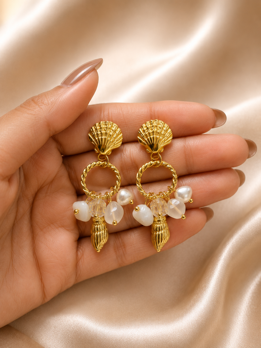 A pair of gold-toned shell and pearl drop earrings held in a person's hand against a soft beige satin fabric background. statement earrings for women, shell earrings, pearl drop earrings, beach inspired earrings, gold statement earrings, anti tarnish earrings, waterproof earrings, 18kt gold plated earrings, stainless steel earrings, coastal jewellery, lightweight earrings, comfort jewellery, vacation earrings, luxury earrings for women, Ashaarya earrings