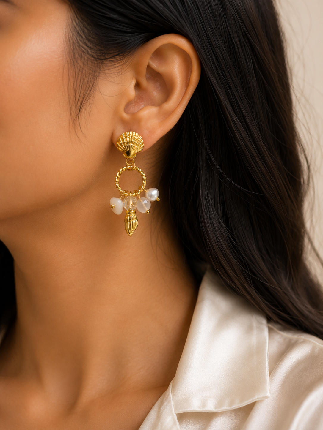 A close-up of a woman wearing a gold dangle earring with white pearl-like beads and a scalloped top, shown against her ear and dark hair. statement earrings for women, shell earrings, pearl drop earrings, beach inspired earrings, gold statement earrings, anti tarnish earrings, waterproof earrings, 18kt gold plated earrings, stainless steel earrings, coastal jewellery, lightweight earrings, comfort jewellery, vacation earrings, luxury earrings for women, Ashaarya earrings