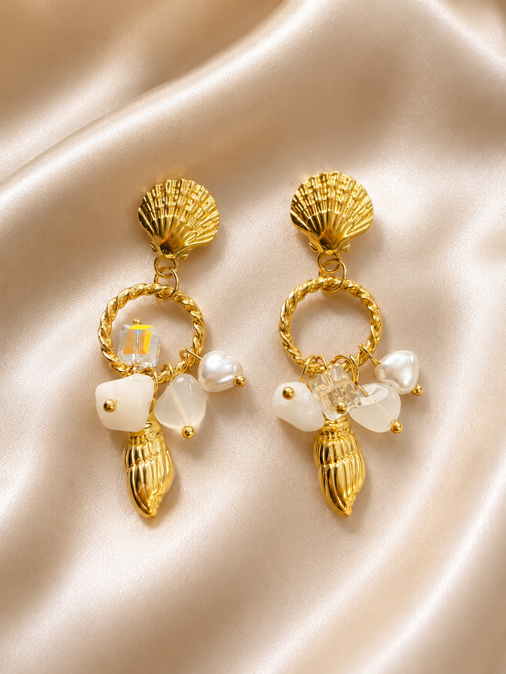 A pair of gold-toned shell-shaped drop earrings with twisted circular hoops, white faux pearls, and clear crystal accents, displayed on a beige satin fabric background. 