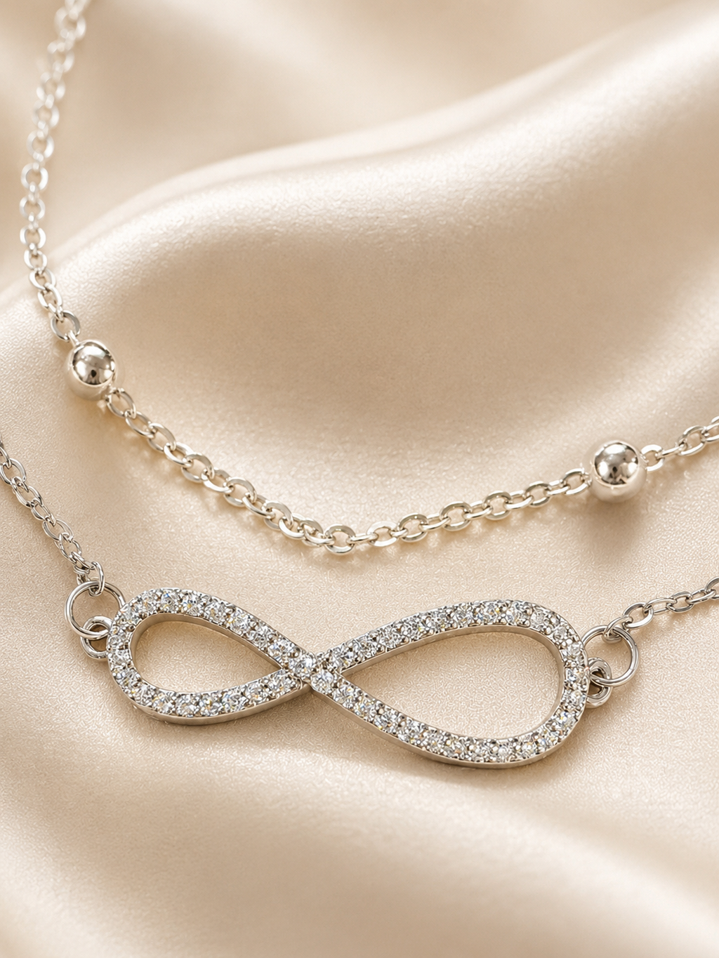 A silver infinity necklace paved with small clear stones, displayed on draped champagne satin fabric.
